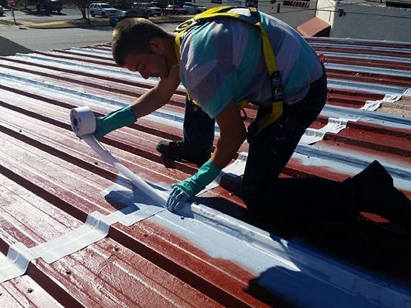 Metal-Roof-Leak-Repair-Canton-GA-Georgia-2 | Rise Residential Roofing Construction worker on a metal roof applying white sealant with a roller, wearing a safety harness and gloves.